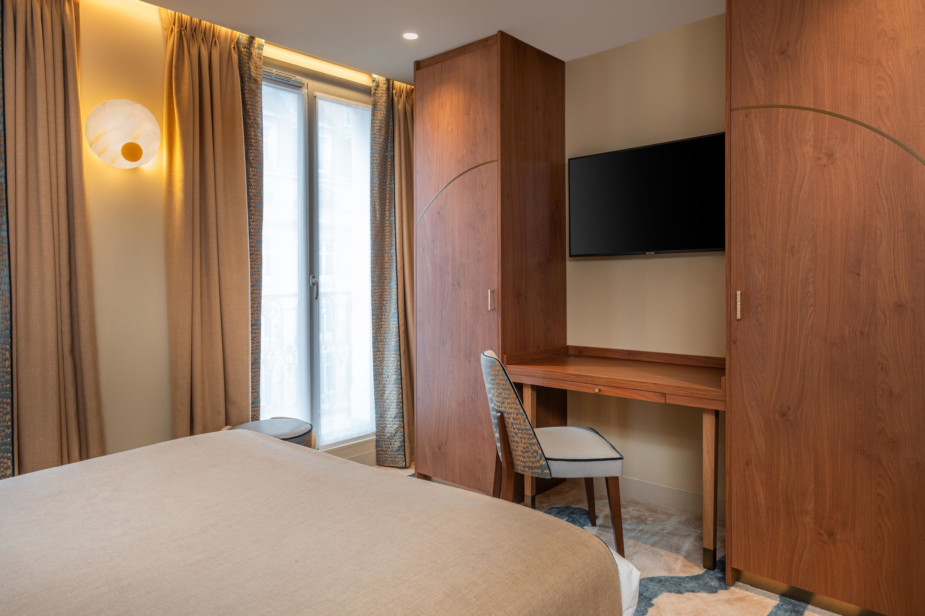 Ett hotellrum med en säng, ett skrivbord, en stol, en tv och ett stort fönster med gardiner.