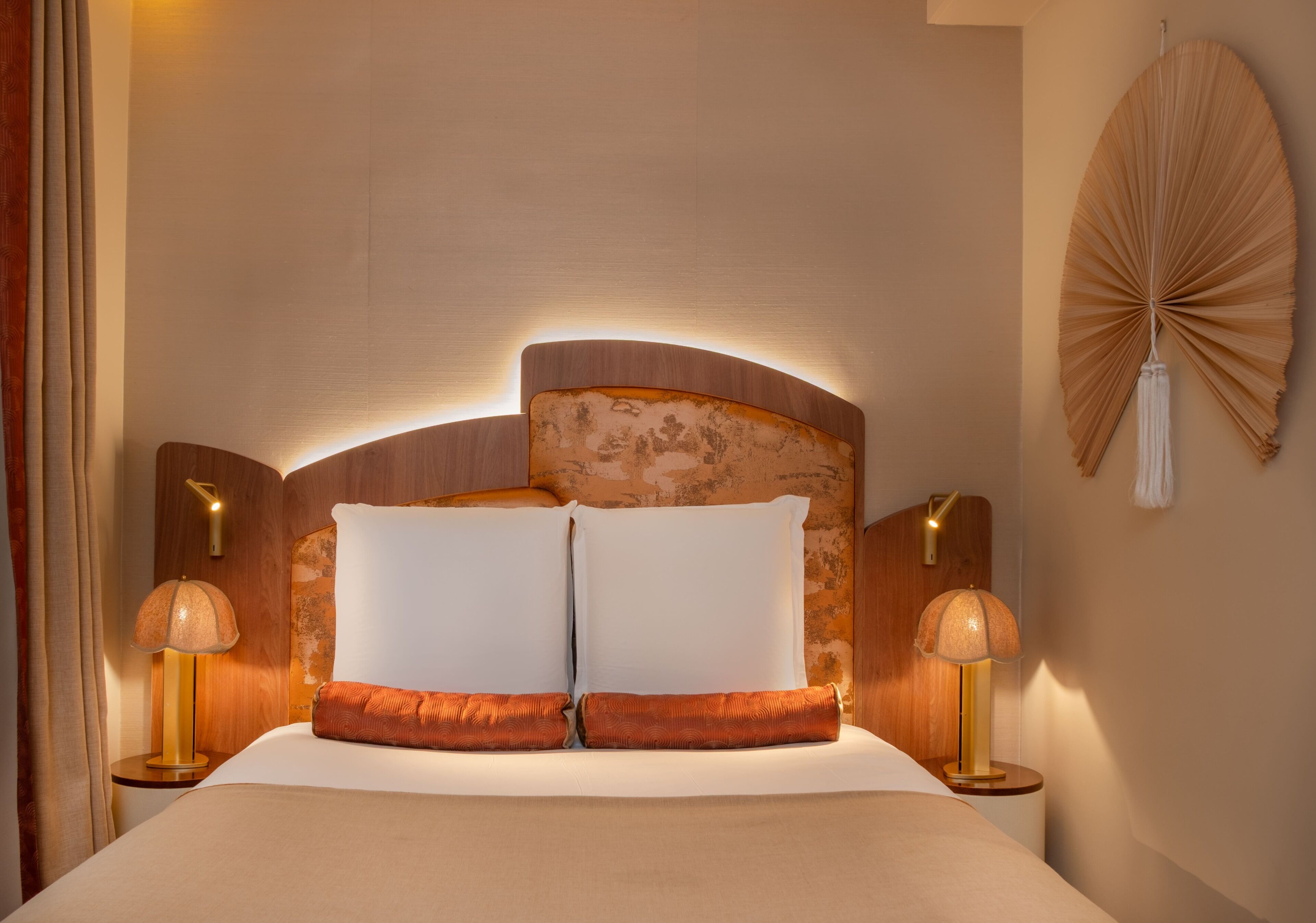 A hotel room with a bed, two bedside lamps, a wall-mounted fan, and a wooden headboard.