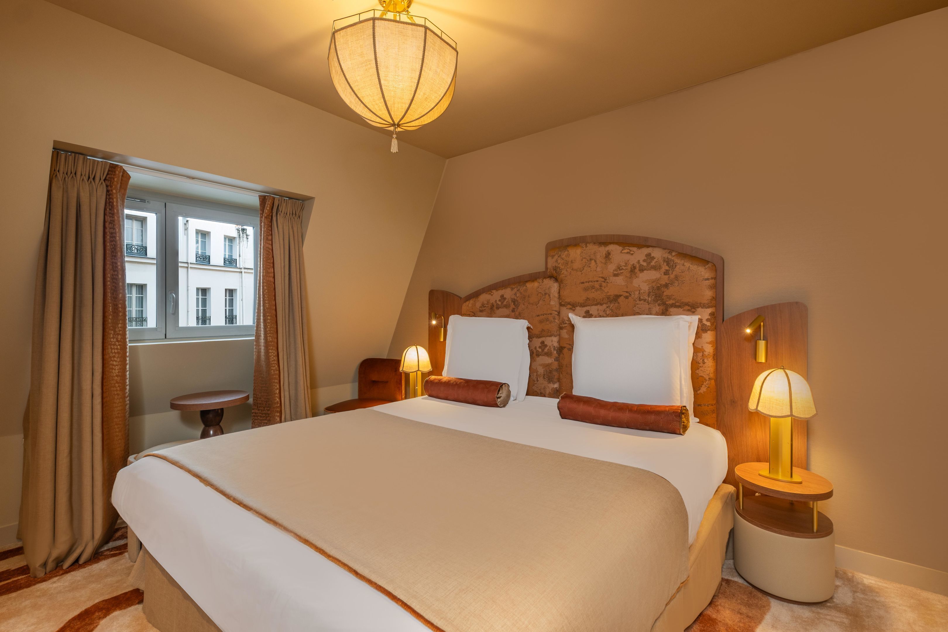 A hotel room with a large bed, two bedside tables with lamps, a window with curtains, and a view of buildings outside.