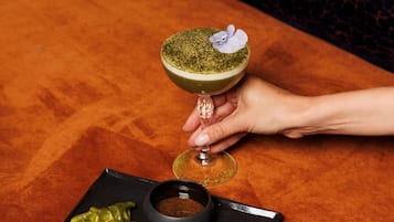 A hand holding a cocktail glass with a flower on top, placed on a black plate with green dumplings and a small bowl of sauce.