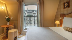Une chambre d’hôtel avec un lit, une petite table sur laquelle est posé un vase de fleurs, une lampe et une vue sur un immeuble par la fenêtre.