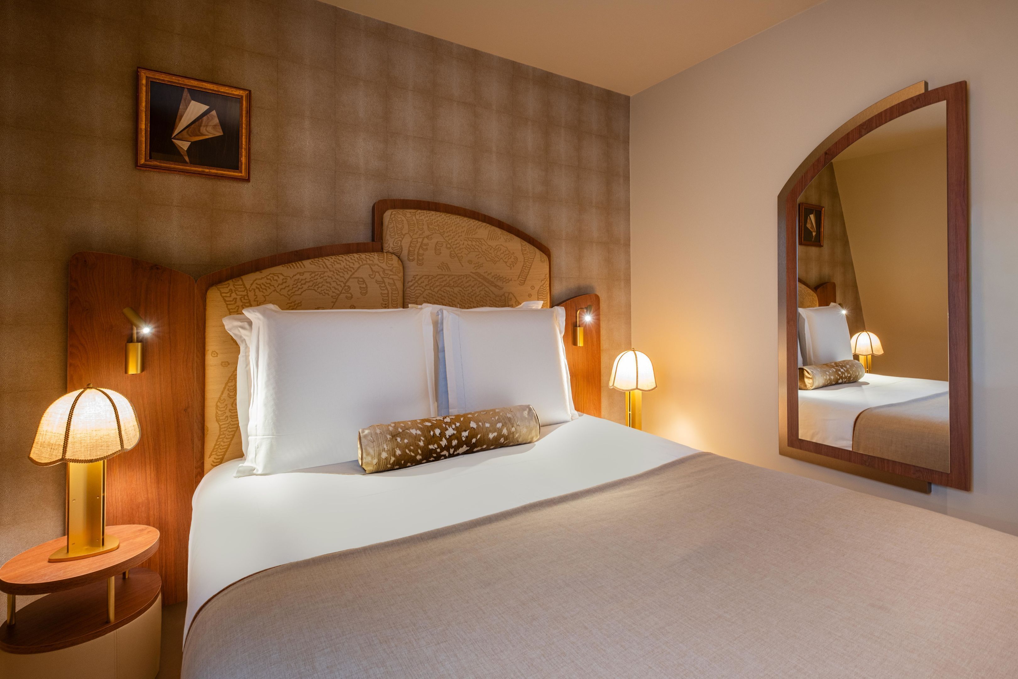 A hotel room with a large bed, two bedside lamps, a wooden headboard, a wall-mounted picture, and a mirror.