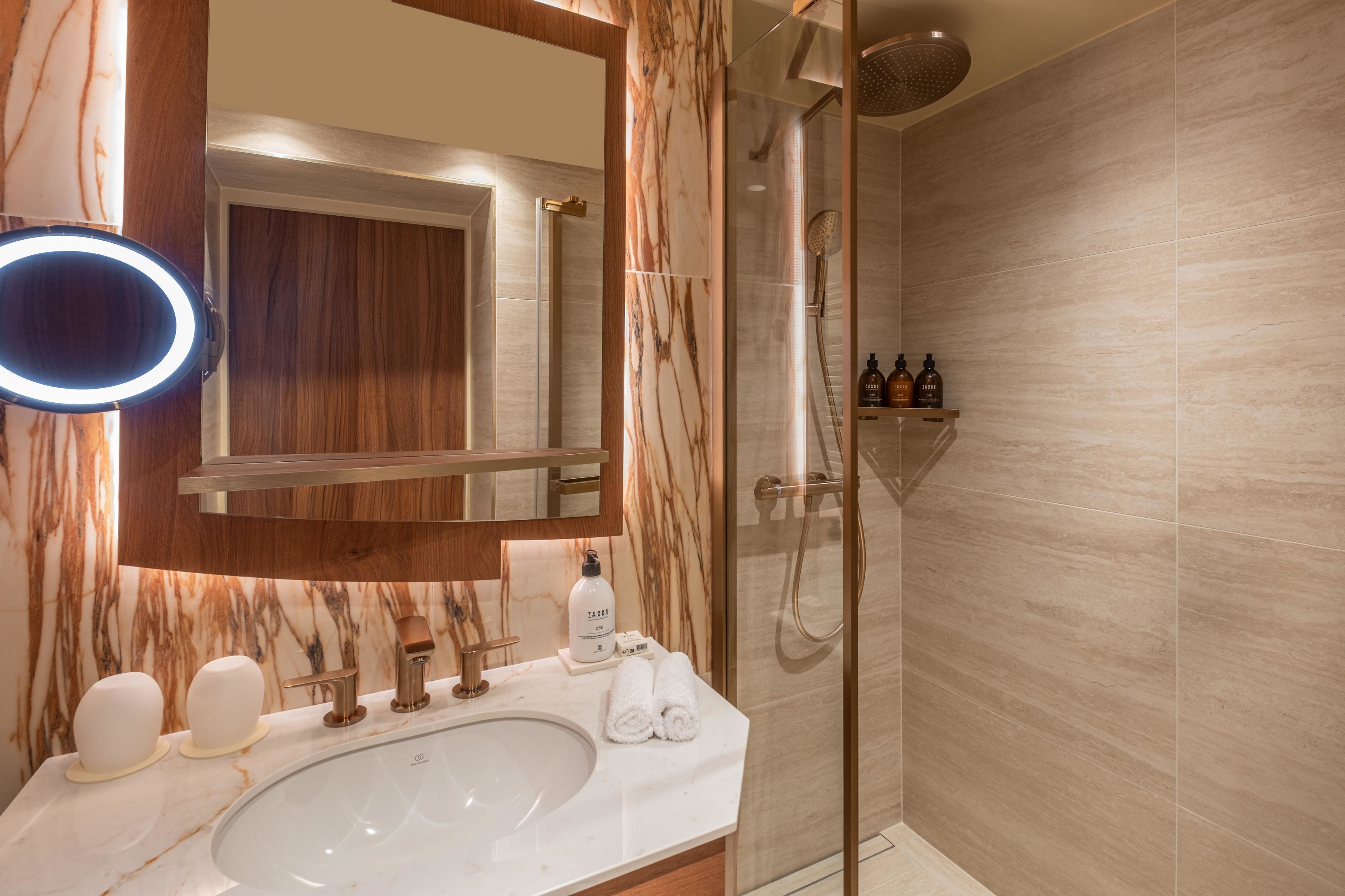 A modern bathroom with a marble vanity, a round mirror with a light, a glass-enclosed shower, and a shelf with toiletries.