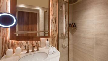 A modern bathroom with a marble vanity, a round mirror with a light, a glass-enclosed shower, and a shelf with toiletries.