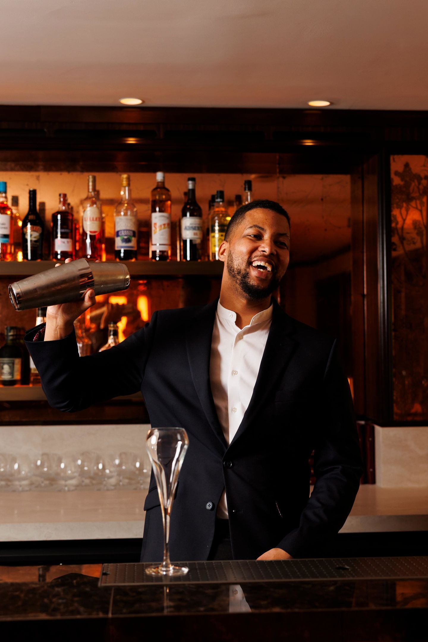 A person in a suit holding a cocktail shaker, standing in front of a bar with various bottles and glasses.