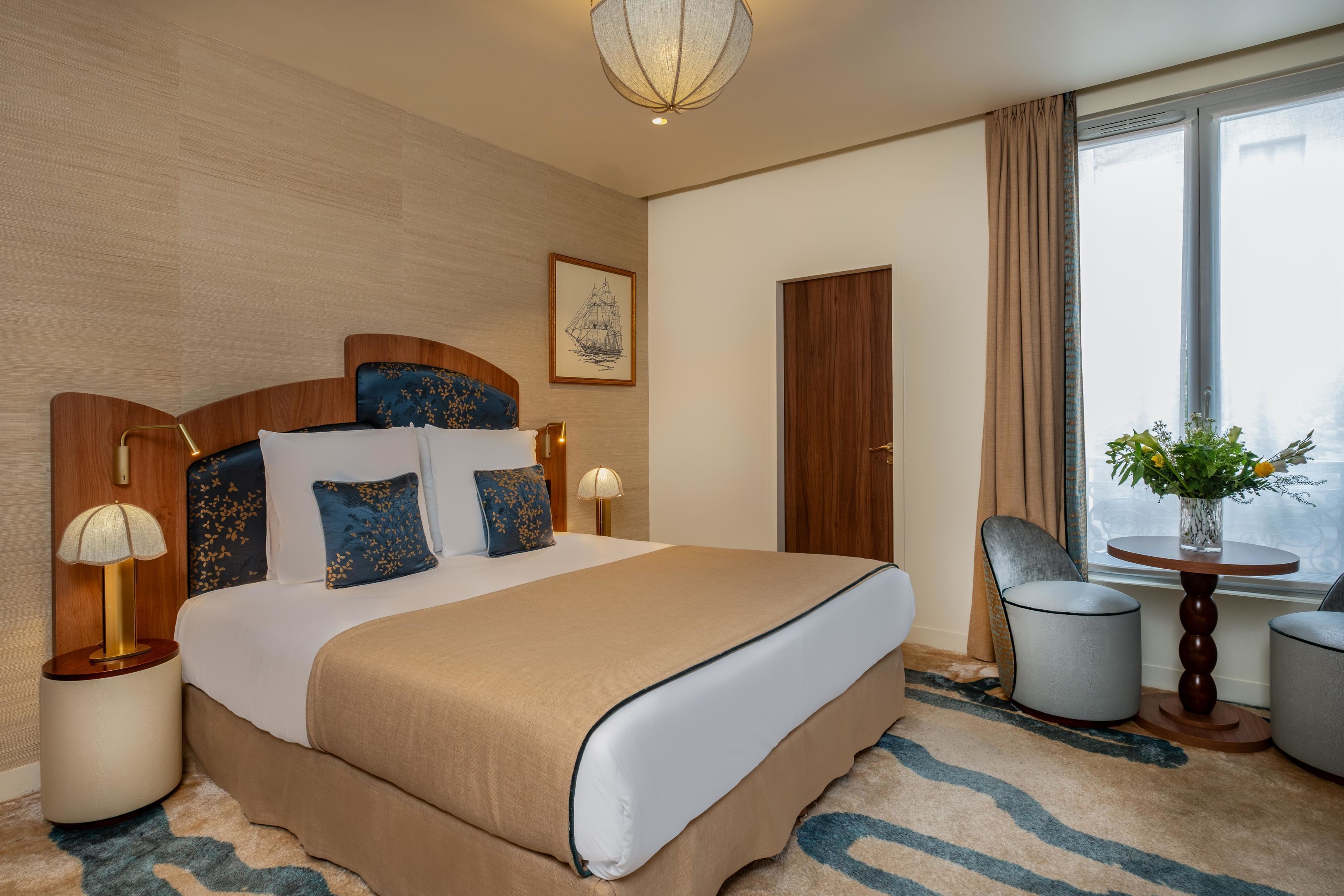 A hotel room with a large bed, bedside lamps, a wooden headboard, a framed picture on the wall, a window with curtains, a chair, a small table, and a vase with flowers.