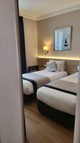 Quarto de hotel com duas camas, uma mesa de cabeceira, um abajur, um quadro e vista para a cidade.