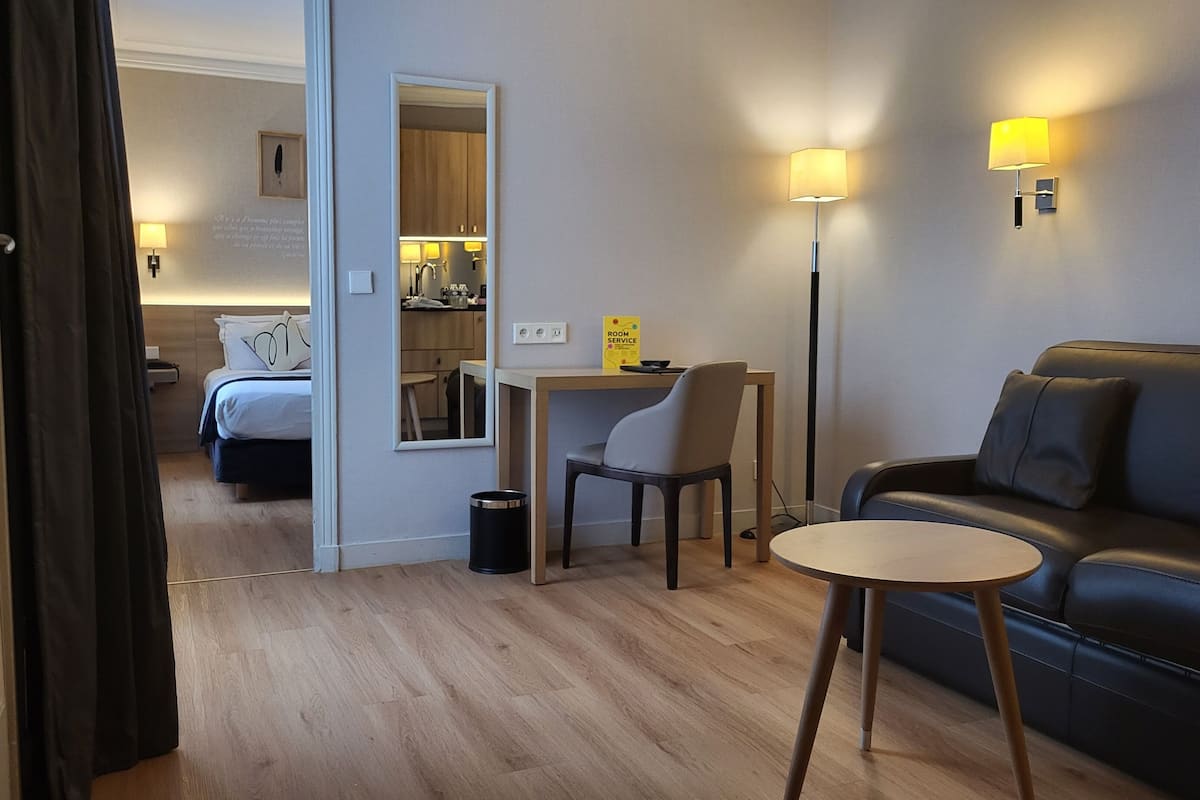 A modern hotel room with a bed, a desk, a chair, a small table, a sofa, and two wall-mounted lamps.