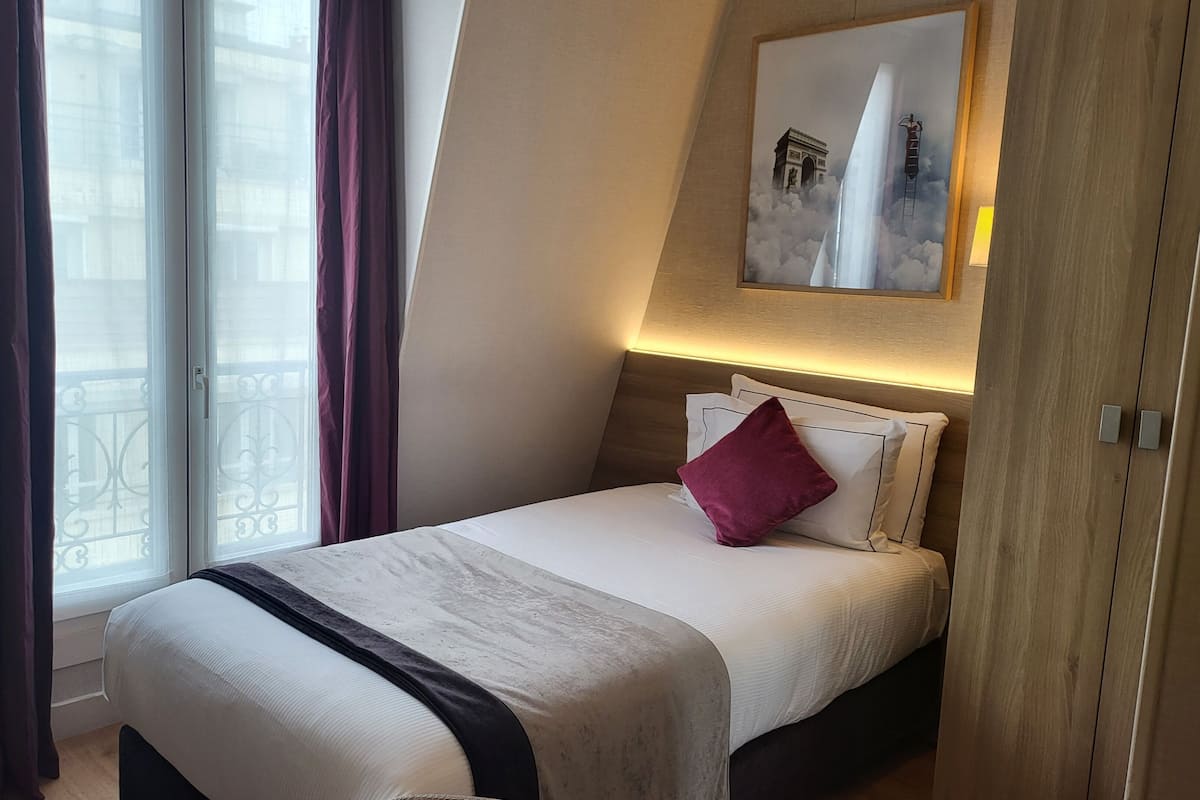 A hotel room with a bed, a nightstand, a window with curtains, and a painting on the wall.