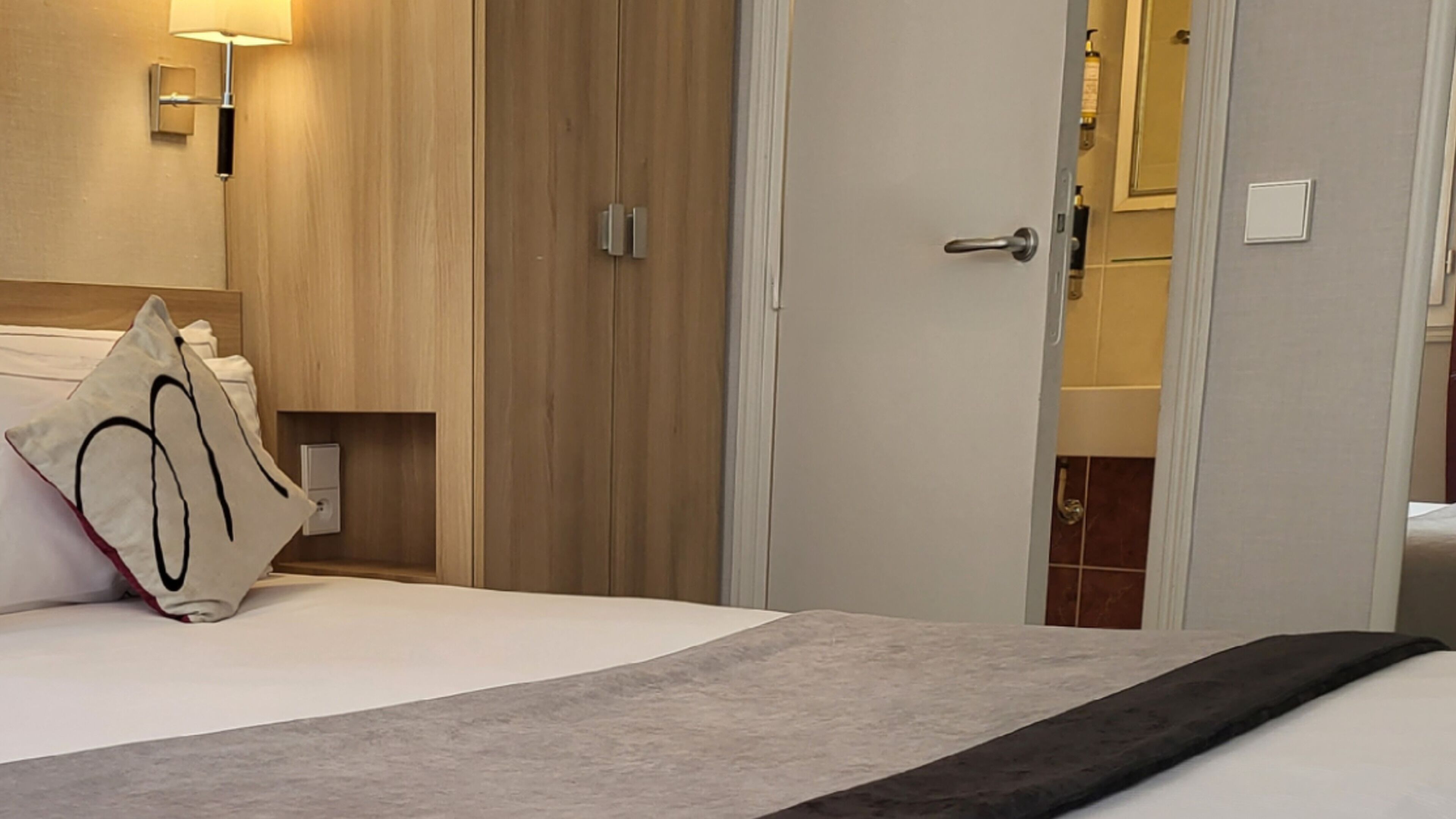 A hotel room with a bed, a wooden wardrobe, a bathroom visible through an open door, and a wall-mounted light fixture.
