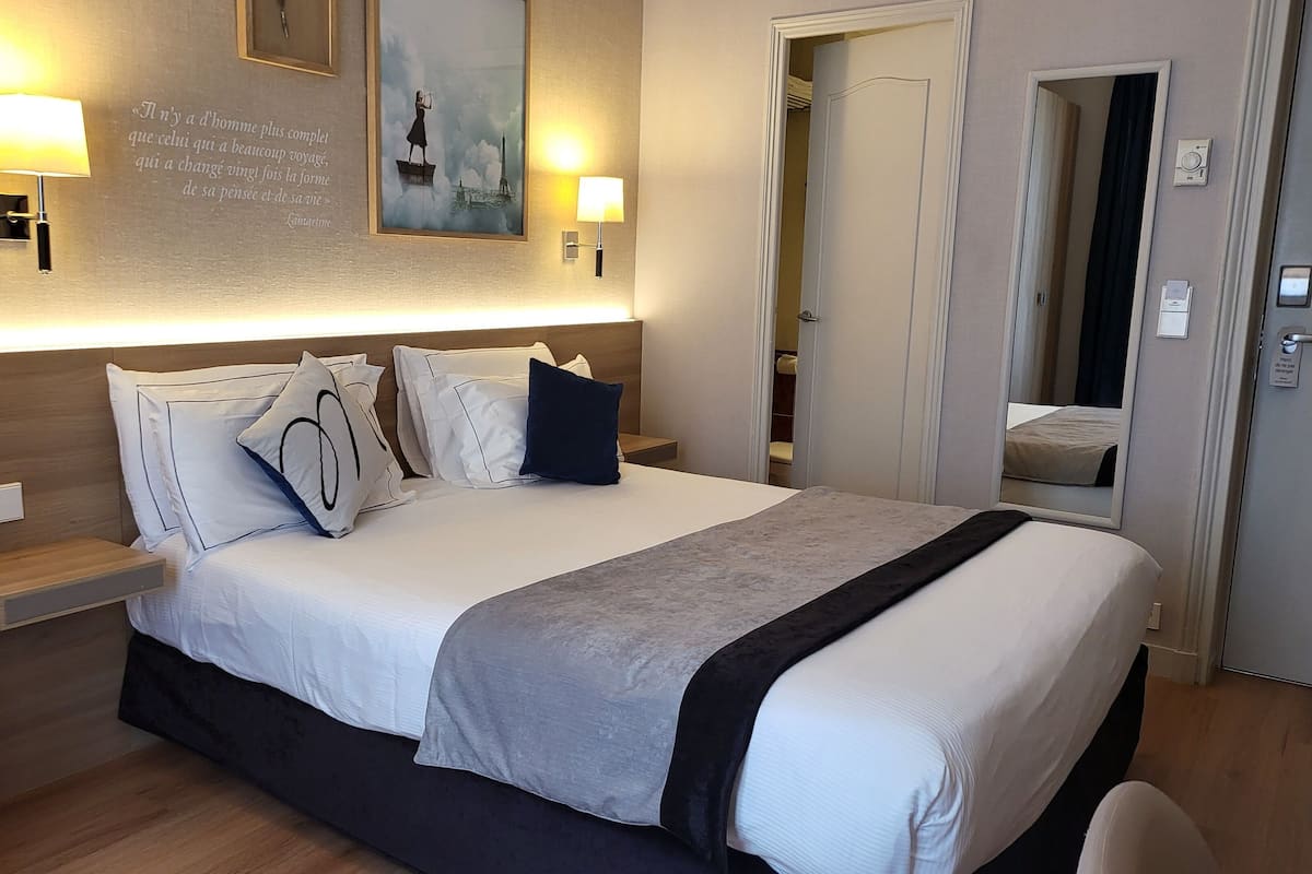 A hotel room with a neatly made bed, bedside lamps, a wall-mounted artwork, and a door leading to another room.