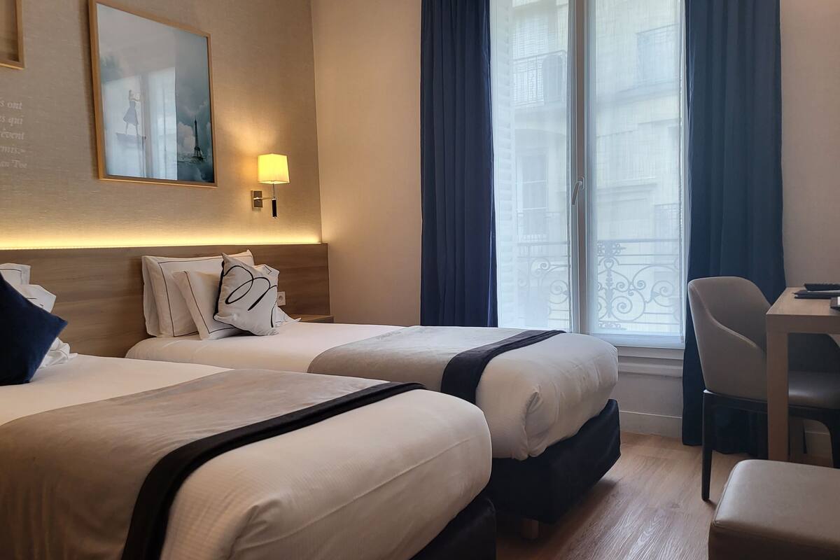 A hotel room with two beds, a desk, and a chair. There is a window with curtains, a lamp, and a framed quote on the wall.