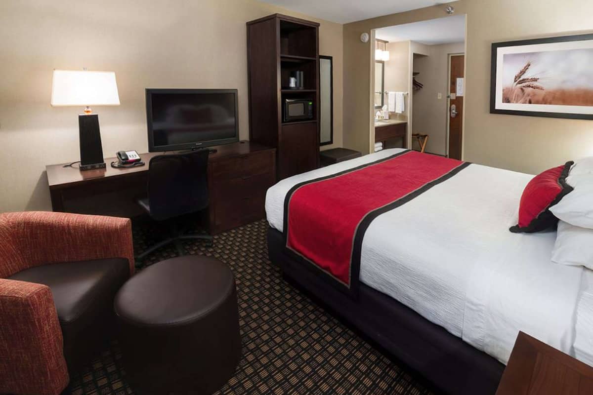 Ein Hotelzimmer mit einem großen Bett, einem Schreibtisch mit Fernseher, einem Sessel, einem kleinen runden Tisch und einer Mikrowelle.