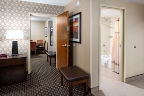 A hotel room with a wooden bench, a desk with a lamp, a framed picture on the wall, and a bathroom visible through an open door. - Best Western Plus Ramkota Hotel (Sioux Falls)