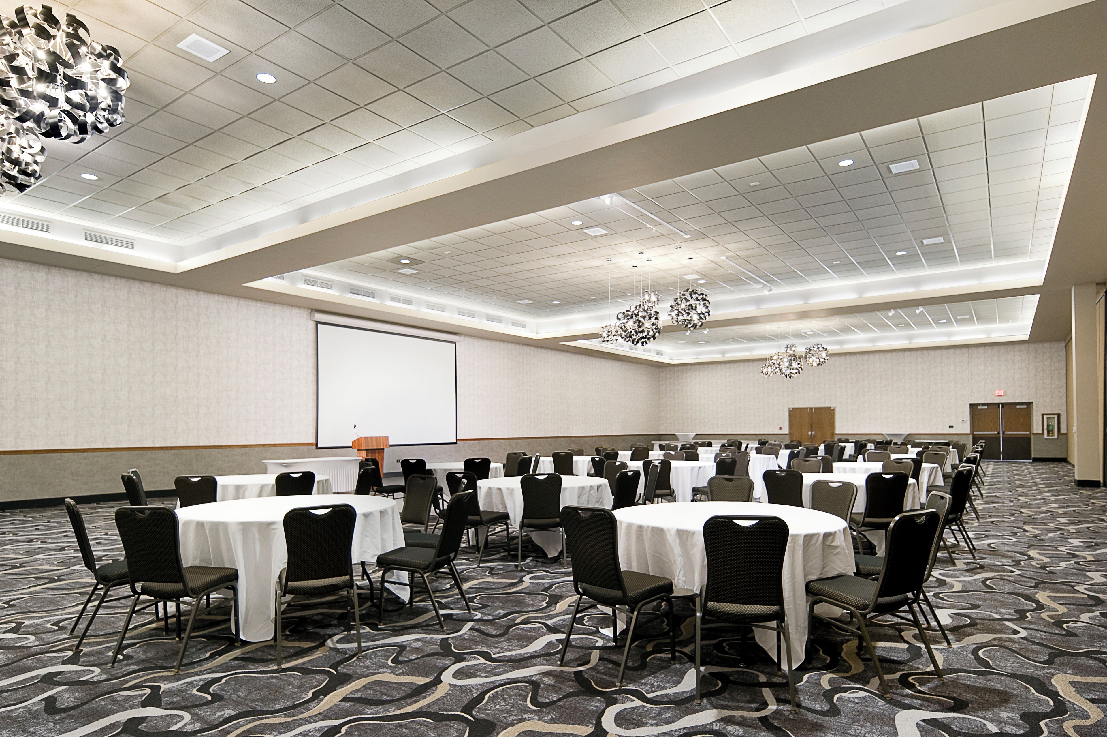 A large conference room with multiple tables and chairs, a projector screen, and a ceiling with recessed lighting.