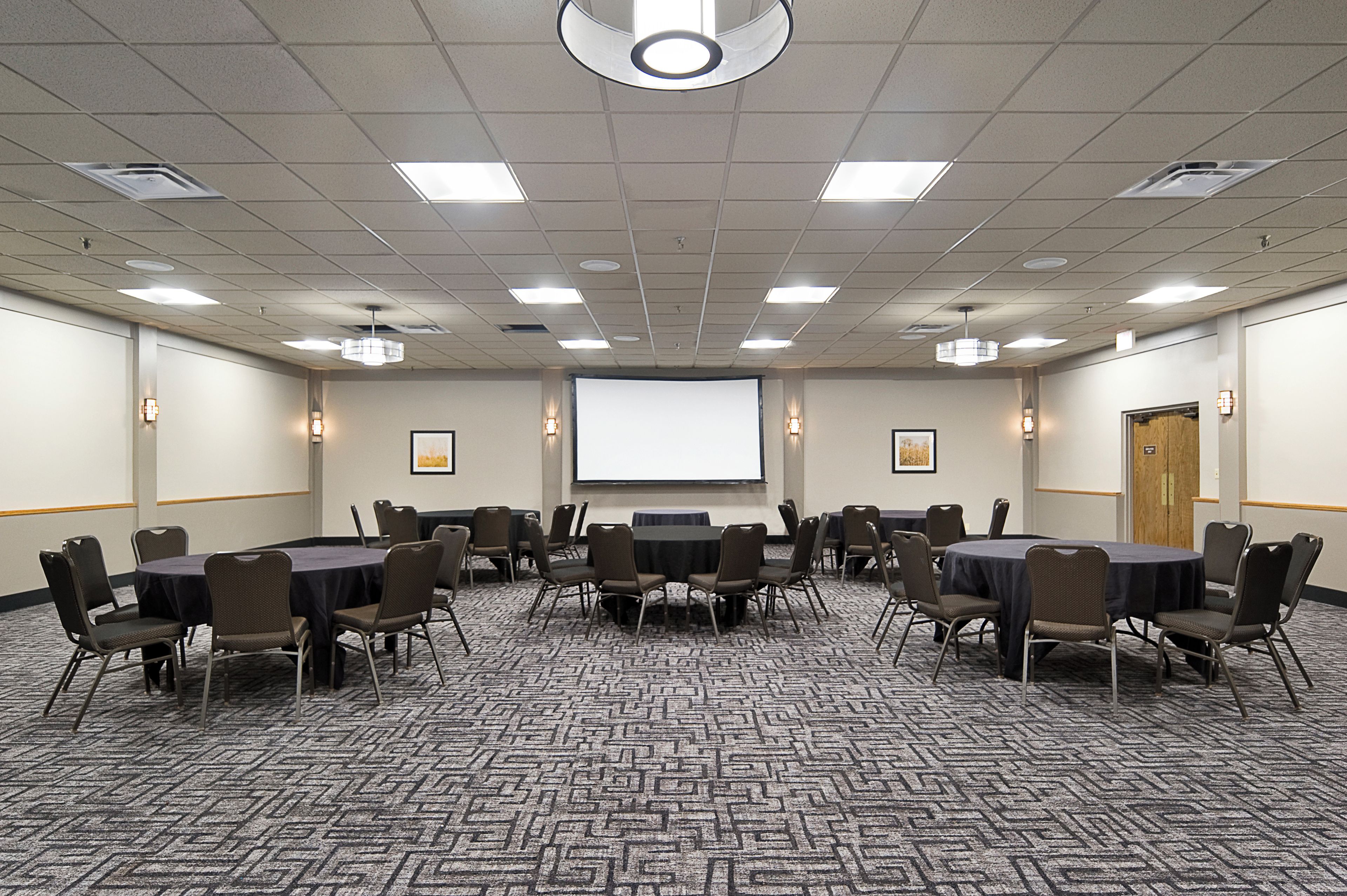 A conference room with multiple tables and chairs, a projector screen, and a carpeted floor.