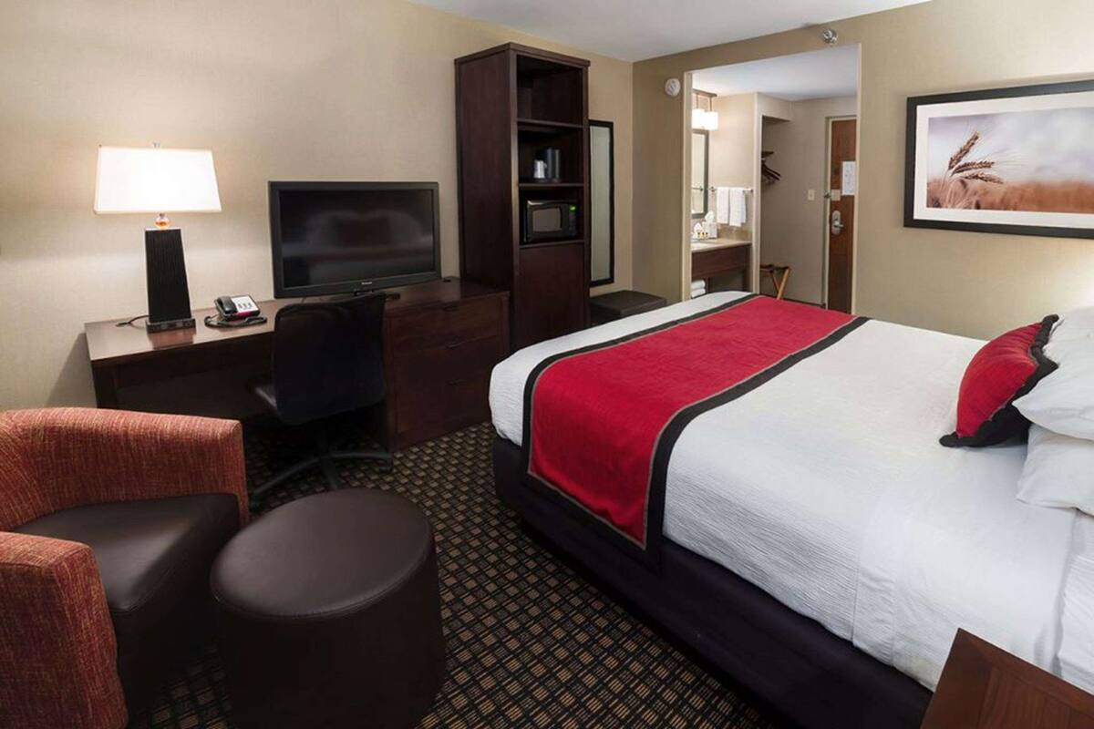 A hotel room with a bed, a desk with a TV, a chair, a small table, and a bookshelf.