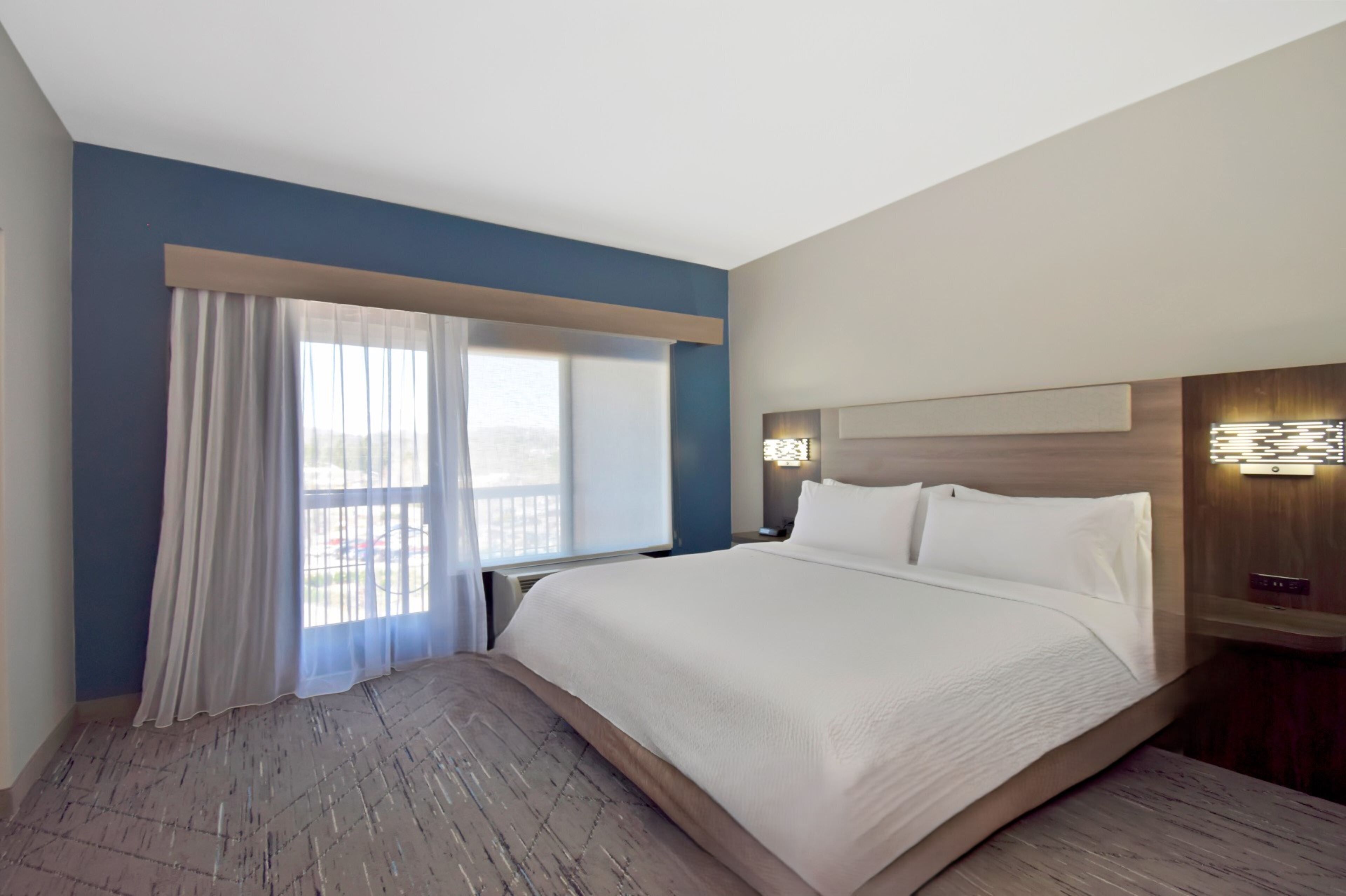 A hotel room with a large bed, a nightstand, a wall-mounted light, and a window with curtains.
