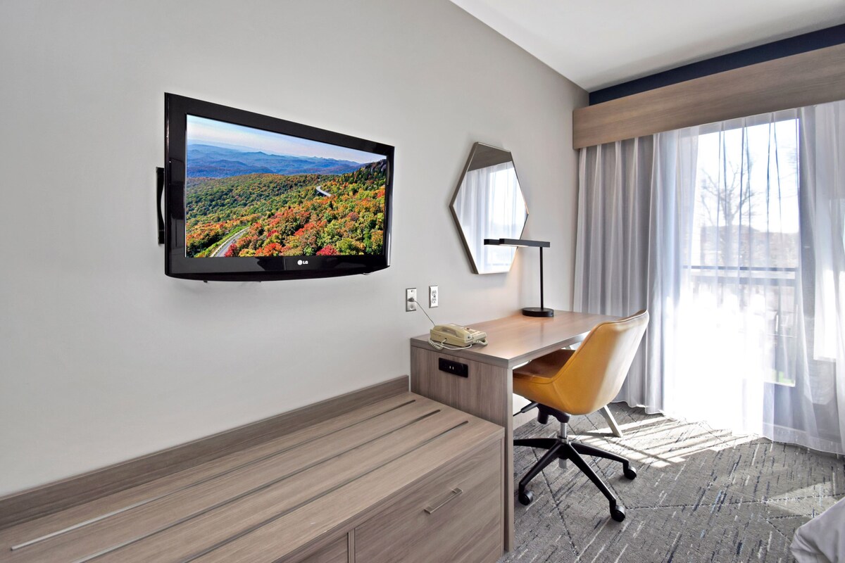 Habitació d'hotel amb televisió de pantalla plana penjada a la paret, escriptori de fusta amb cadira, telèfon i finestra gran amb cortines primes.