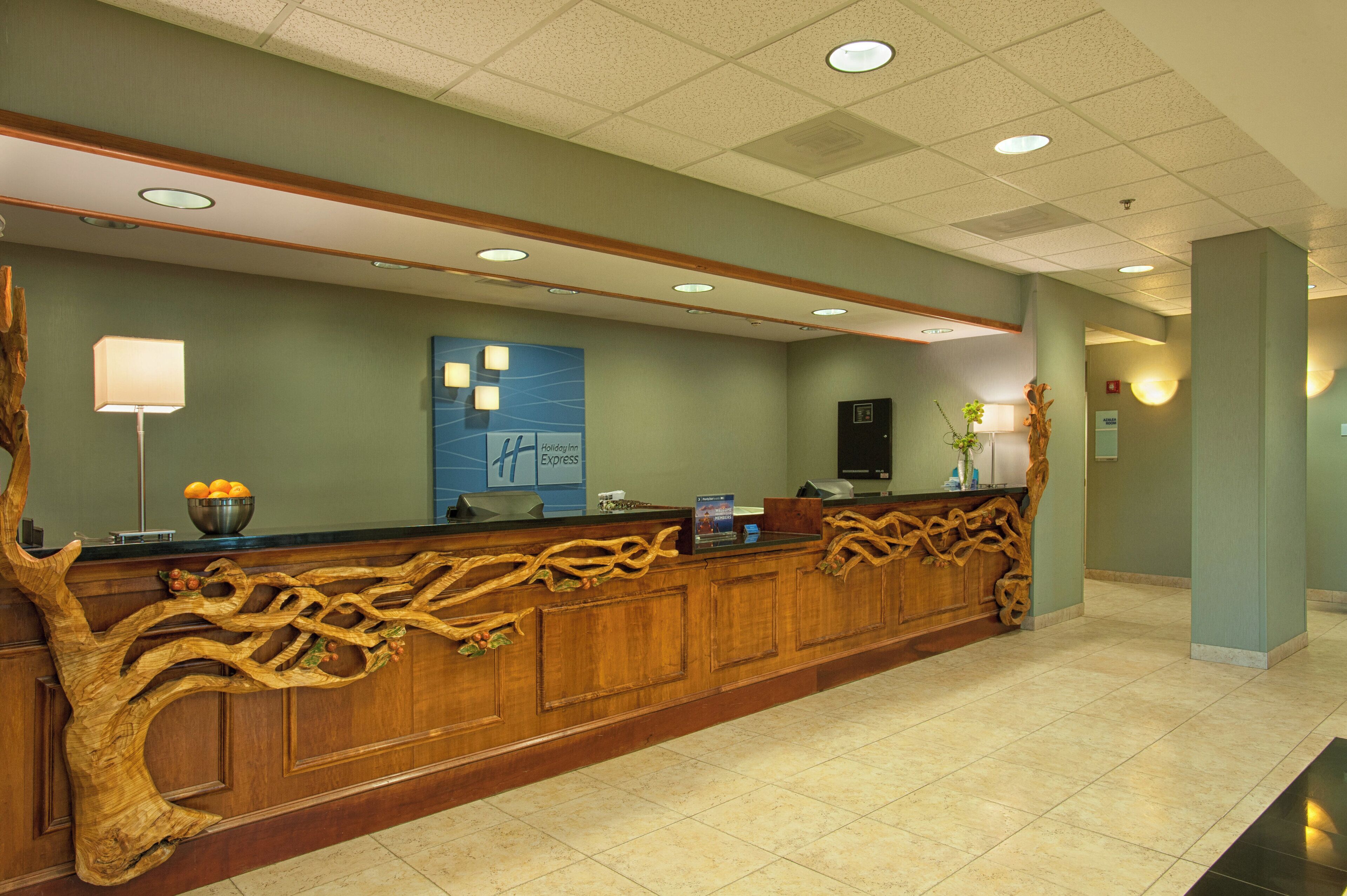 A hotel reception desk with a wooden design, a blue wall with a Holiday Inn Express sign, and a reception area with a flat-screen TV and a vase with flowers.