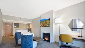 A hotel room with a bed, two chairs, a desk, a fireplace, and a large painting on the wall.