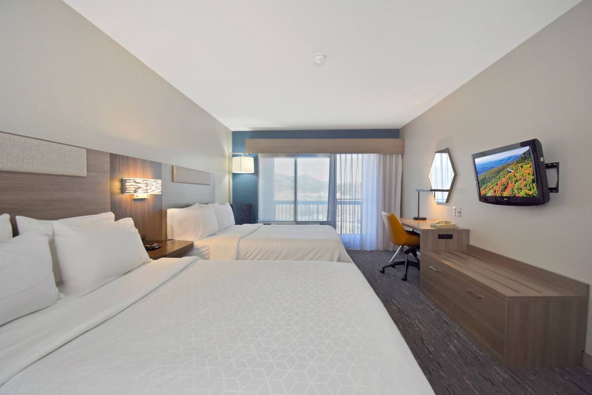 Ein Hotelzimmer mit zwei Betten, einem Schreibtisch, einem Sessel, einem Fernseher und einem Fenster mit Vorhängen.