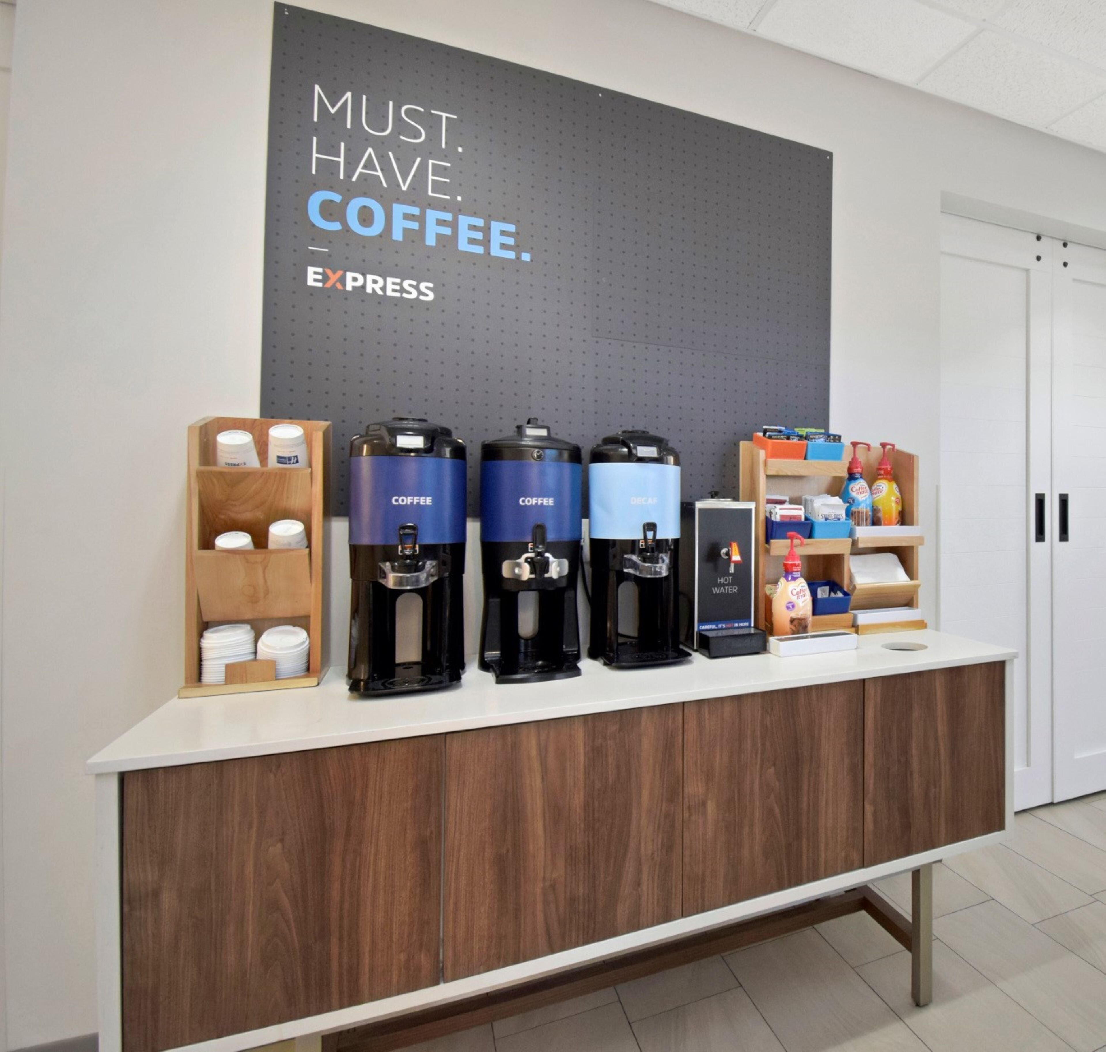 A coffee station with multiple coffee dispensers, a sign saying "MUST HAVE COFFEE. EXPRESS", and a counter with various coffee-related items.