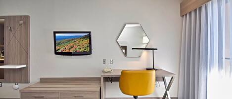 A hotel room with a bed, a desk with a chair, a TV, a mirror, and a window with curtains.