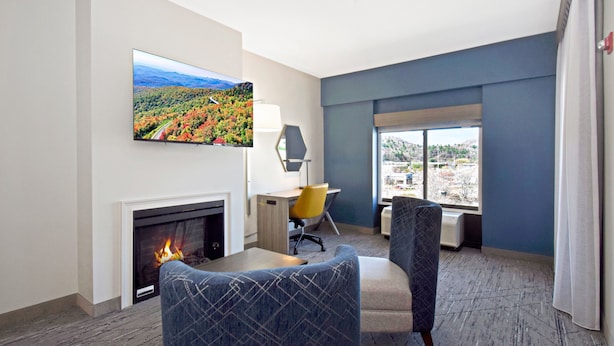 A hotel room with a fireplace, a desk, two chairs, a large window with a city view, and a painting on the wall.