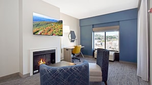 A hotel room with a fireplace, a desk, two chairs, a large window with a city view, and a painting on the wall.