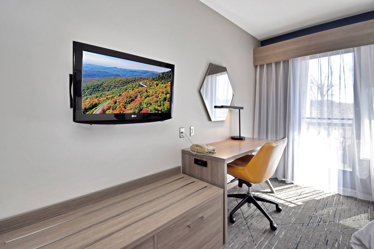 Ein Hotelzimmer mit Flachbildfernseher an der Wand, einem Holztisch mit Stuhl, einem Telefon und einem großen Fenster mit durchsichtigen Vorhängen.