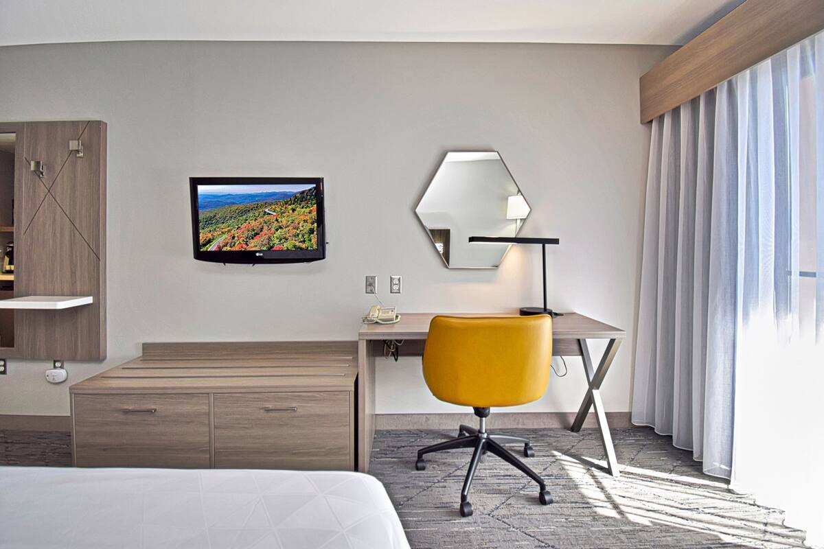 Ein Hotelzimmer mit Bett, Schreibtisch mit Stuhl, Fernseher, Spiegel und einem Fenster mit Vorhängen.