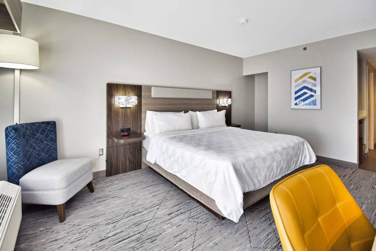 Ein Hotelzimmer mit einem großen Bett, einem Nachttisch mit Lampe, einem Sessel und einem Wandbild.