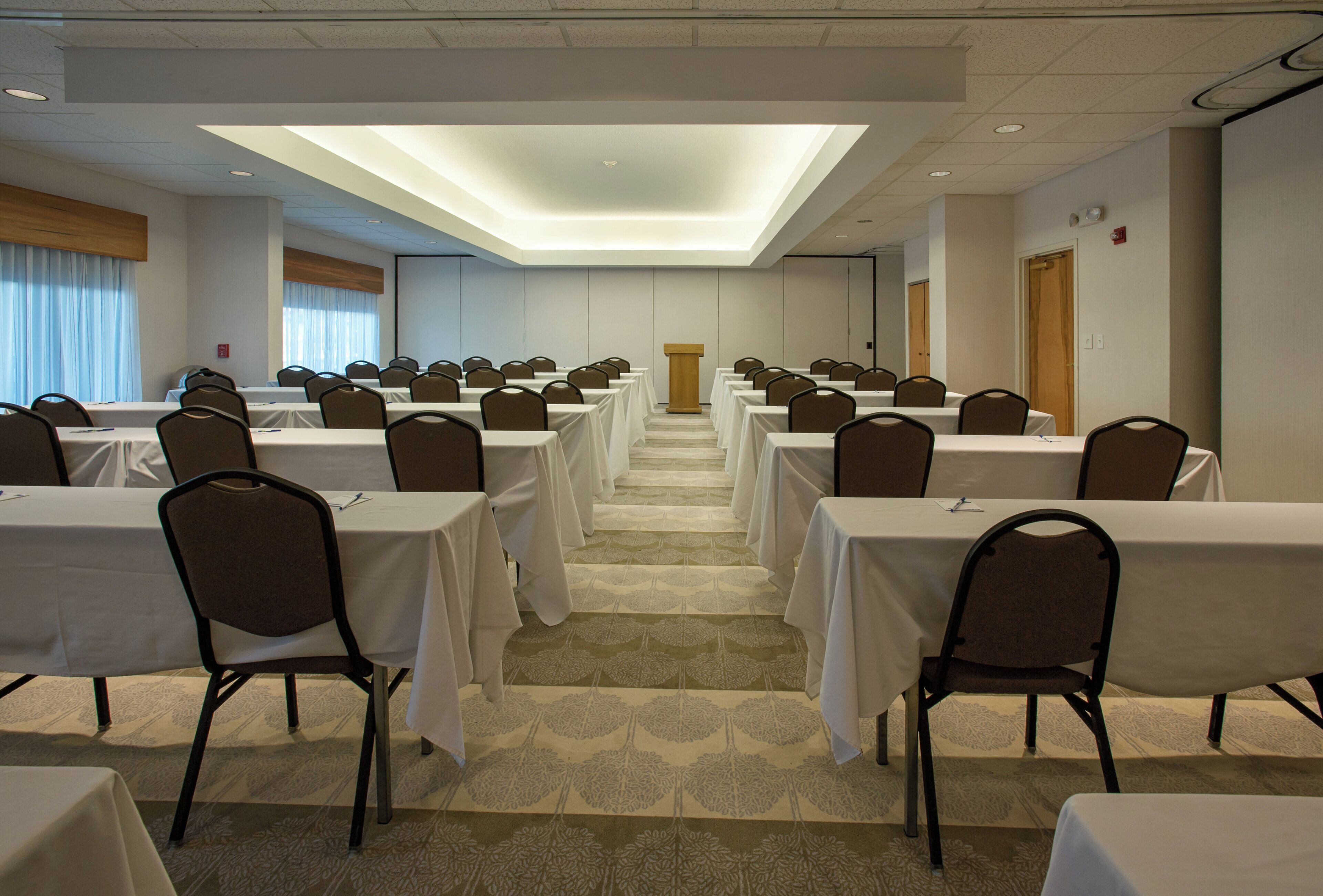 A conference room with rows of tables and chairs, a podium at the front, and a large window with curtains.