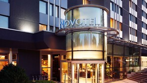 Una entrada de hotel moderna con una puerta giratoria de cristal, una recepción visible y un cartel que dice 'NOVOTEL'.