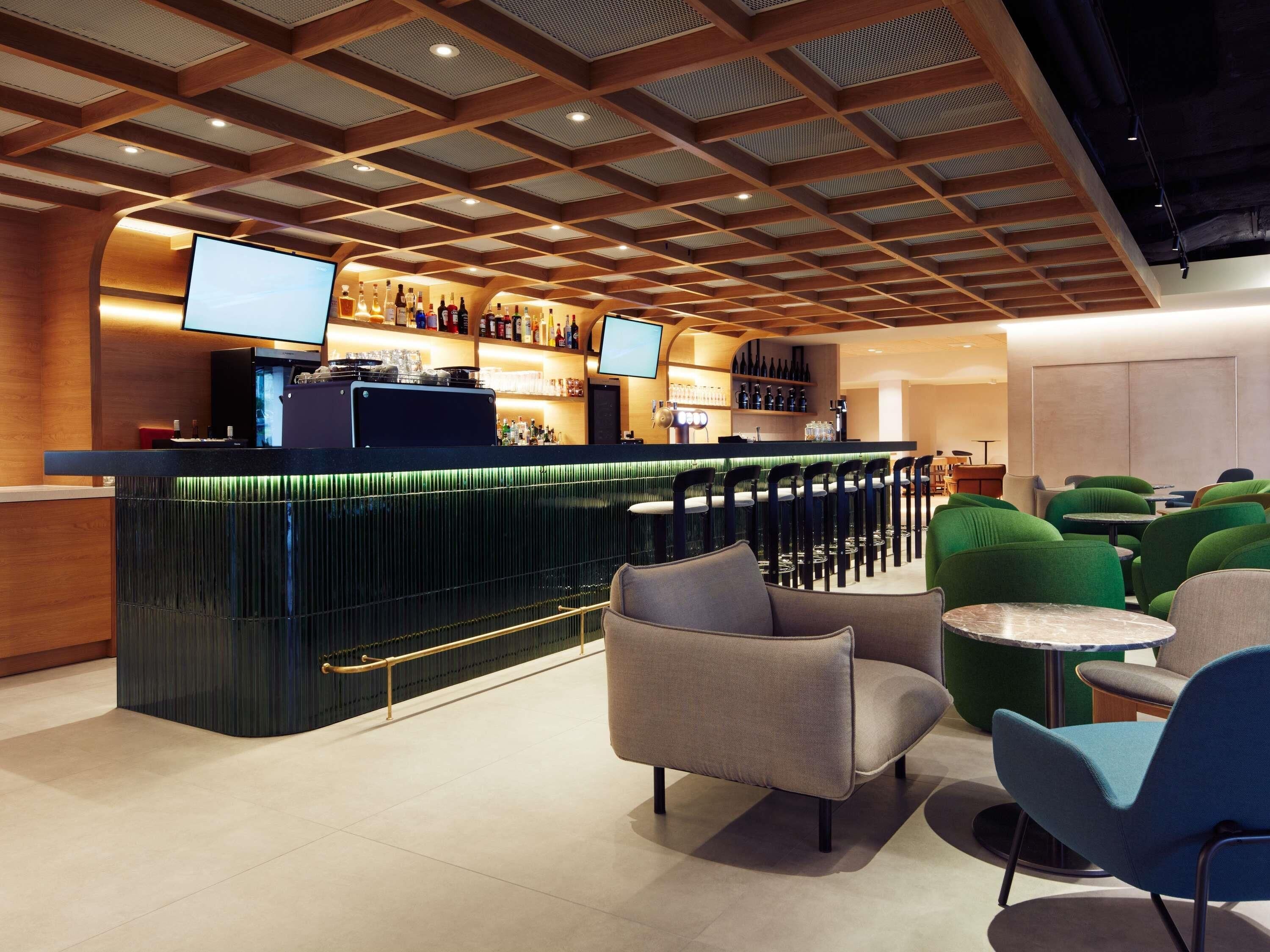 A modern bar area with a wooden ceiling, bar counter, and seating with green and grey chairs.