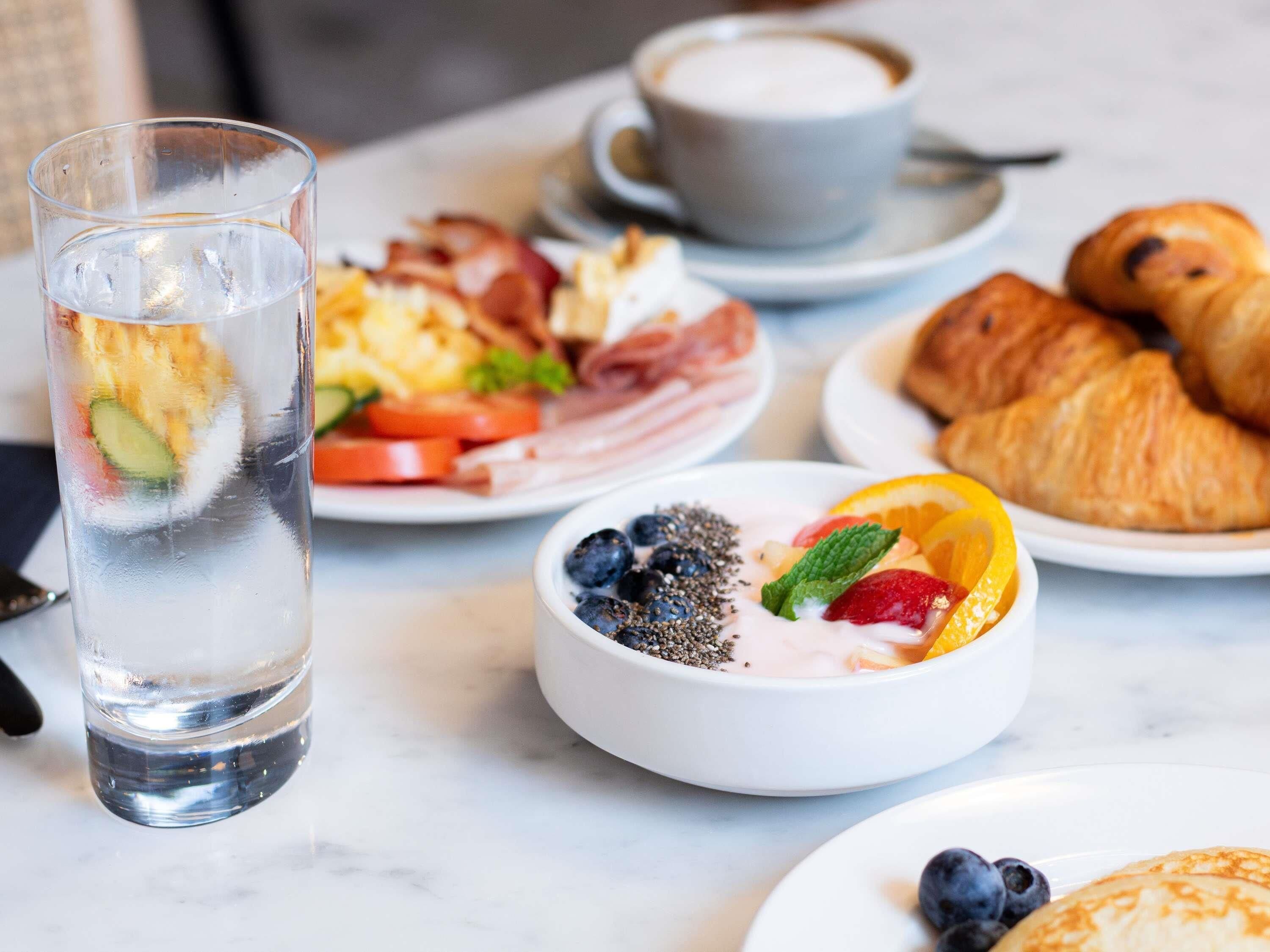 A breakfast setting with a glass of water, croissants, a bowl of yogurt with fruit, and a plate of eggs and bacon.