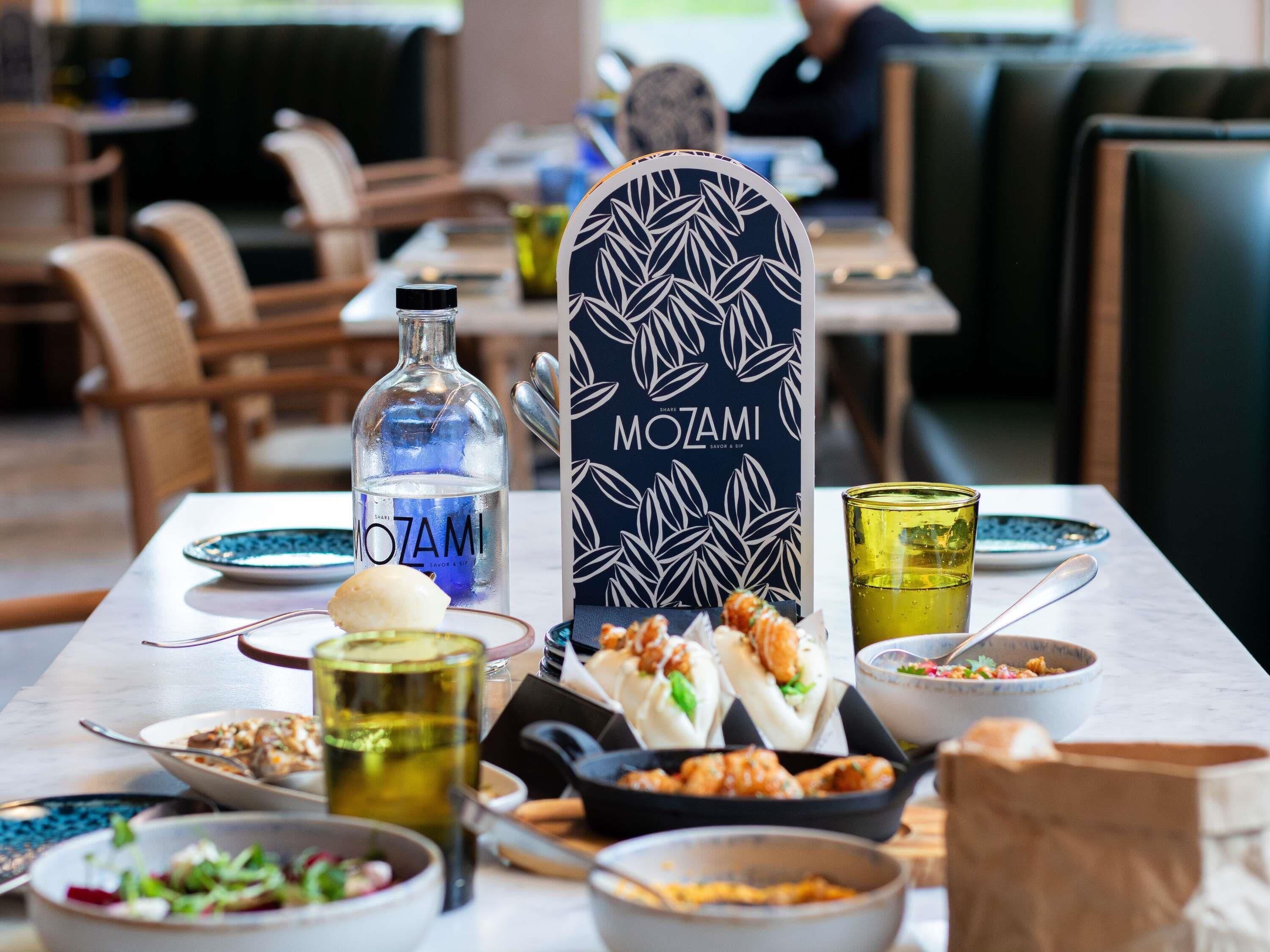 Una mesa de comedor con una botella de MoZAMI, un cartel negro con un diseño de hojas, y diversos platos y bebidas.