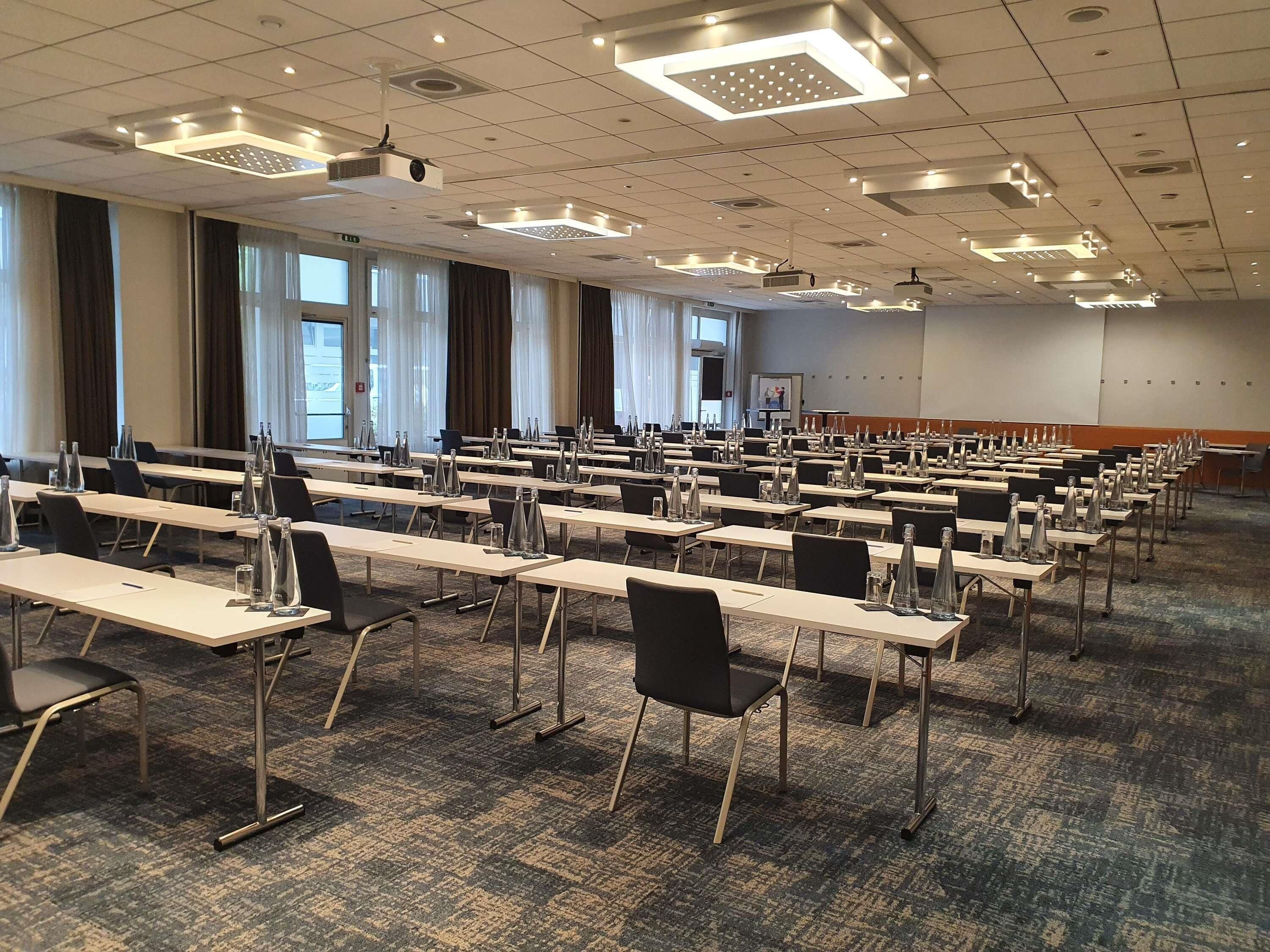 Une salle de conférence avec des rangées de tables, de chaises et un écran de projection.