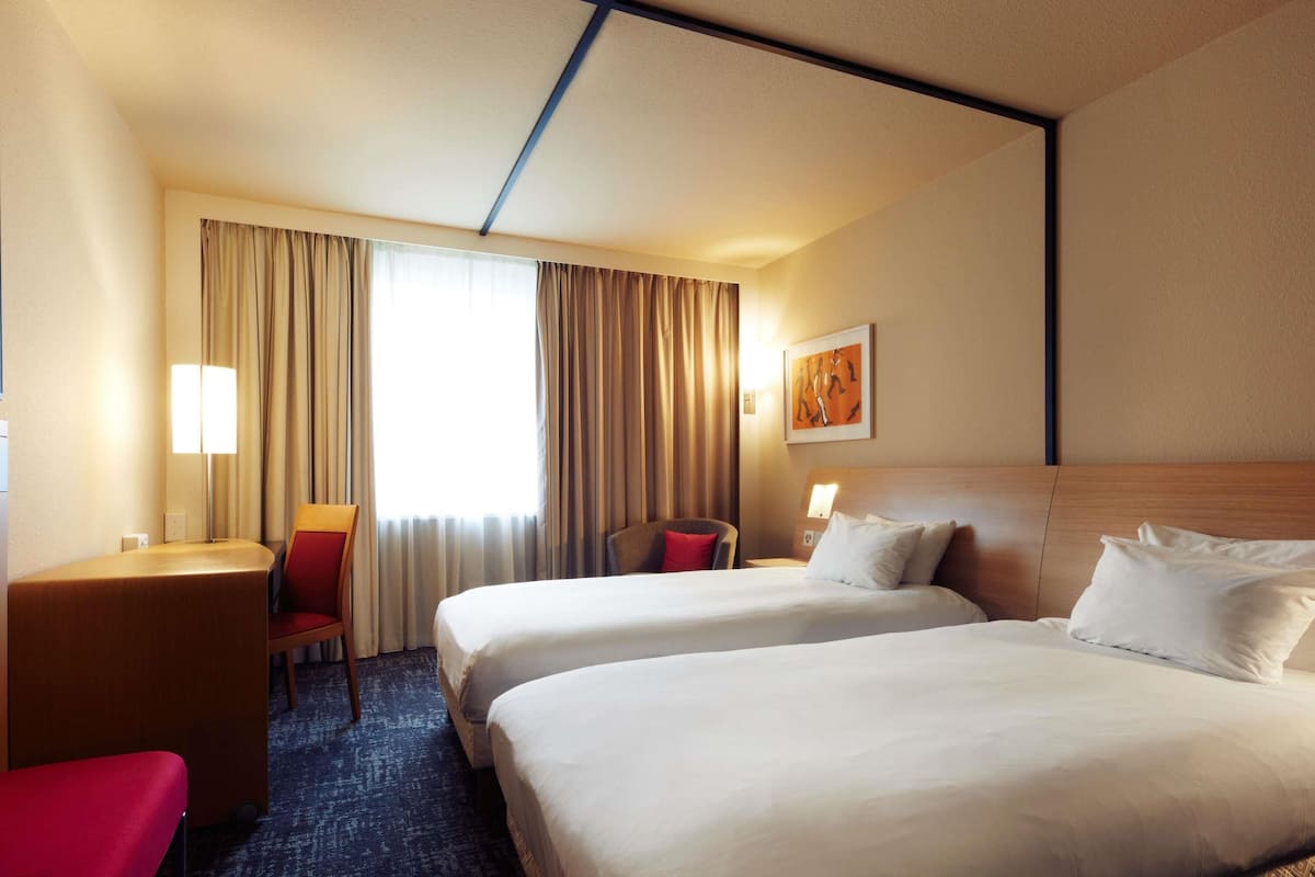 A hotel room with two beds, a desk, a chair, and a large window with curtains.