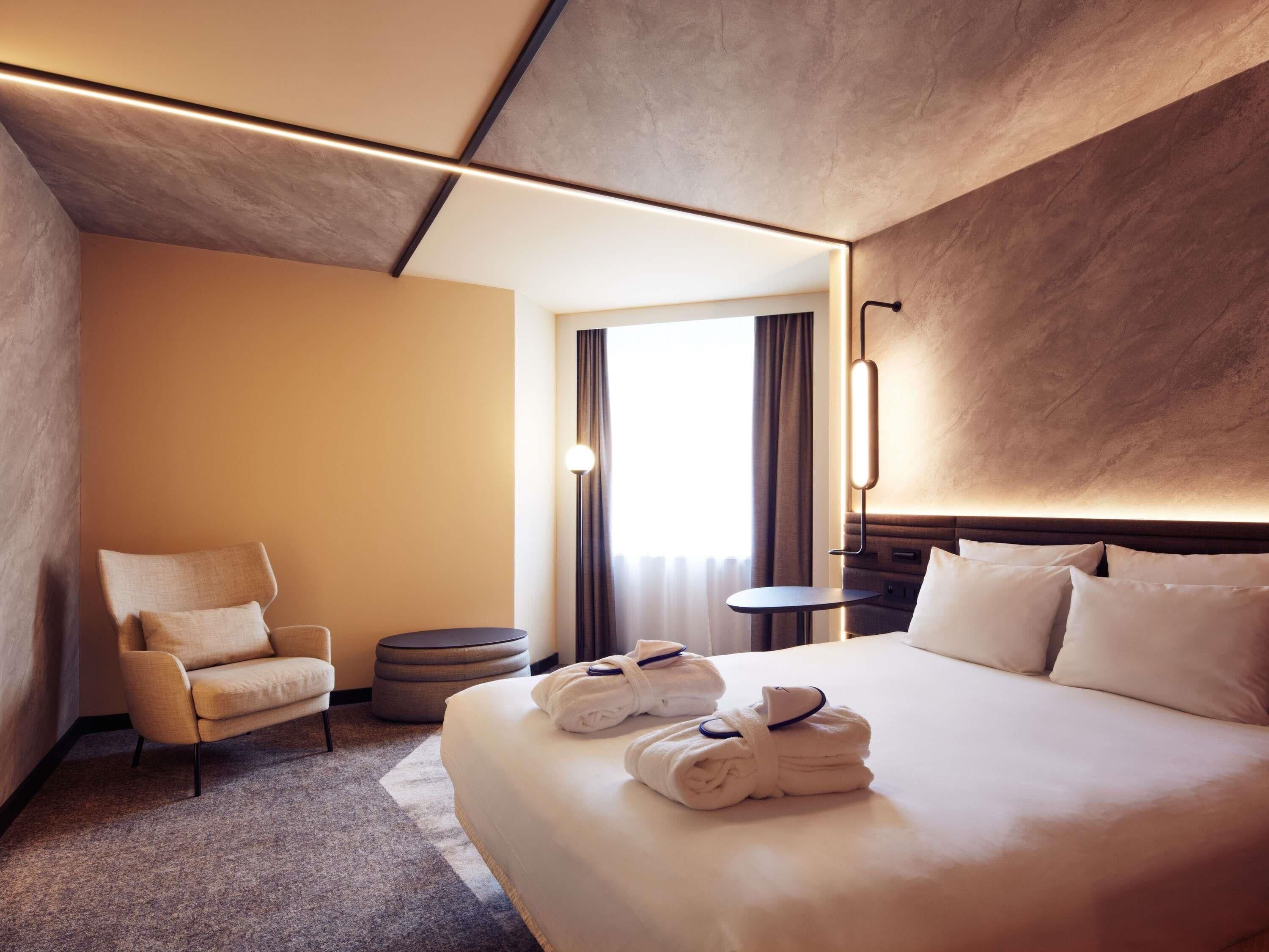 A hotel room with a bed, a chair, a small table, and a round ottoman.