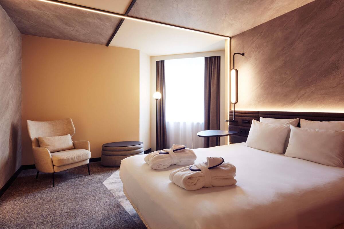 A hotel room with a bed, a chair, a small table, and a round ottoman.