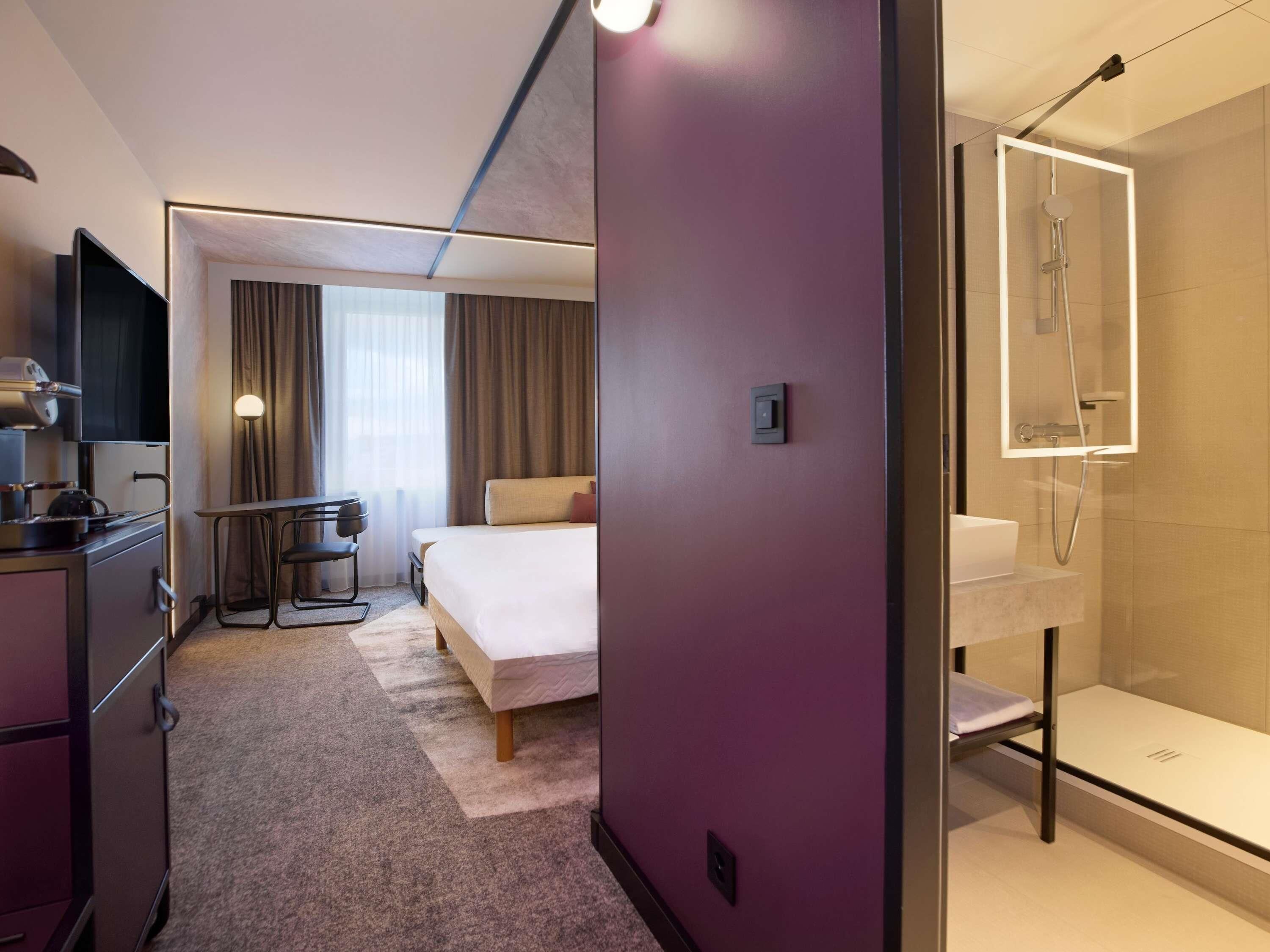 Una habitación de hotel moderna con una cama, un escritorio con silla, televisor, ducha y baño.