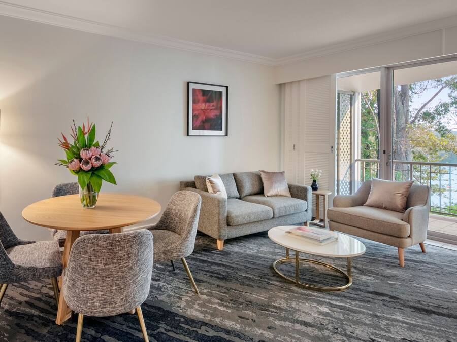 Een moderne woonkamer met een ronde houten tafel, beklede grijze stoelen, een grijze bank en een beige fauteuil. Er is een groot raam met uitzicht op bomen en een balkon.
