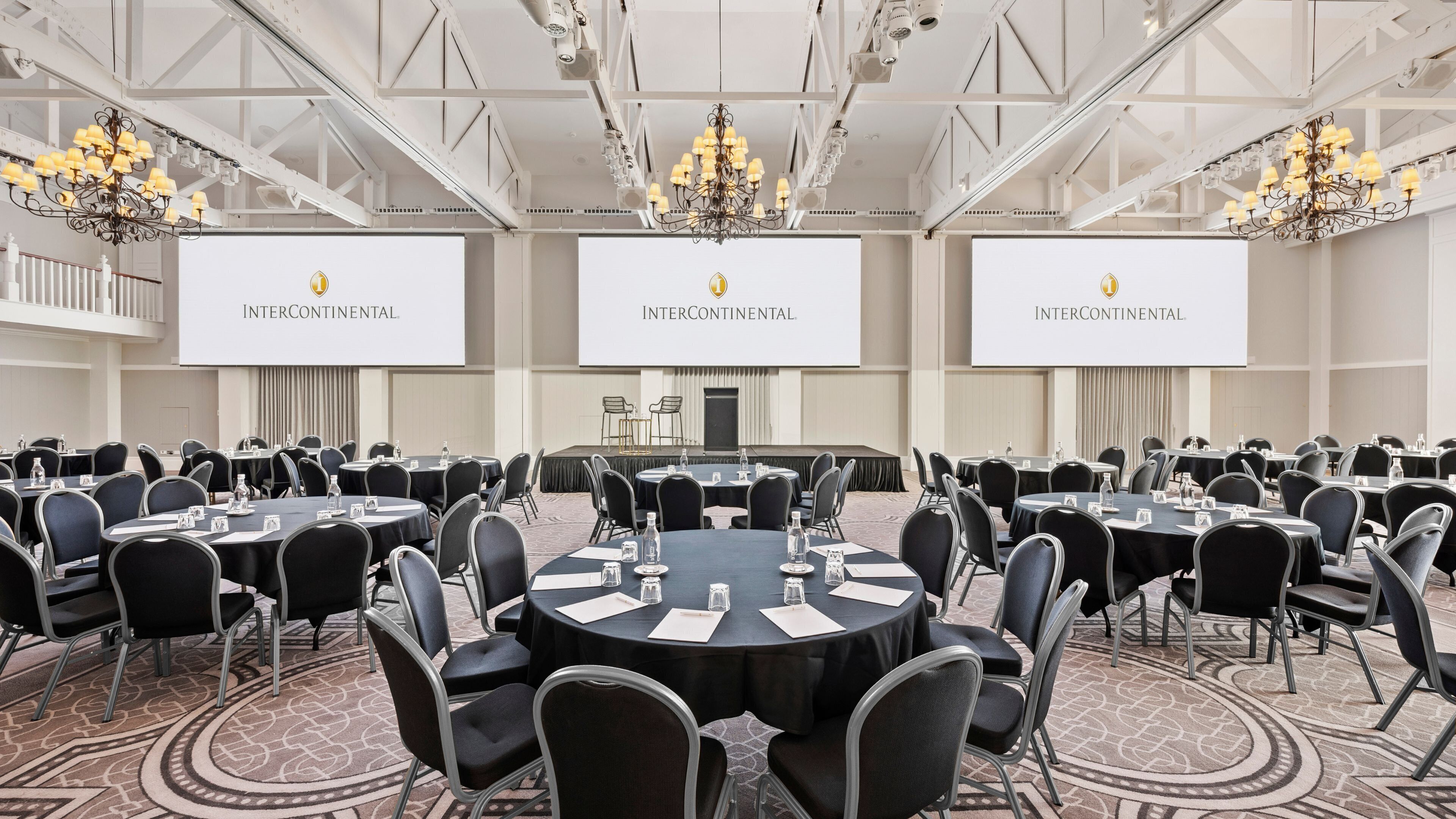 A conference room with multiple tables and chairs, large screens displaying the InterContinental logo, and chandeliers.