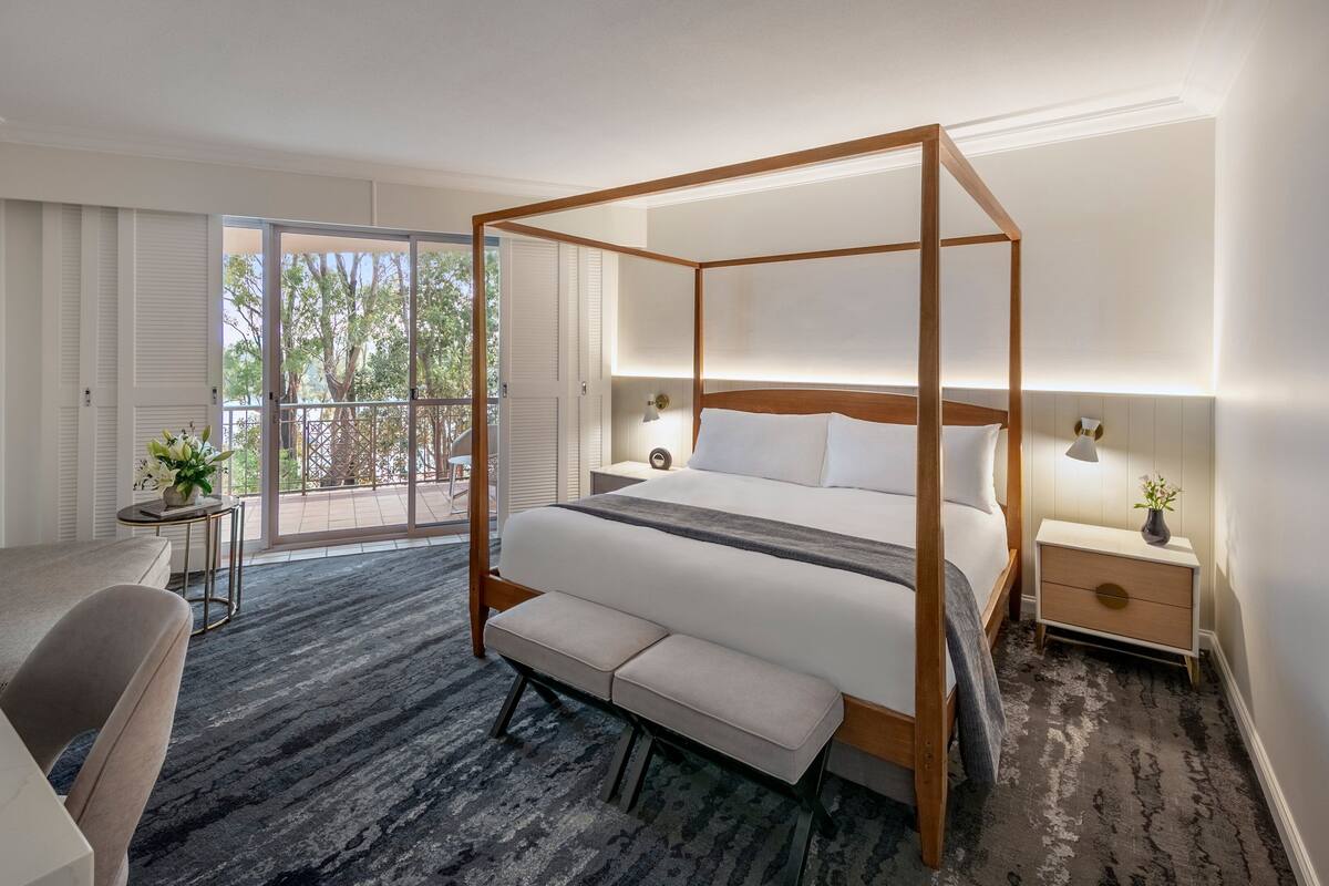 Une chambre d’hôtel moderne avec un lit à baldaquin, un canapé, une petite table et un balcon offrant une vue sur les arbres.