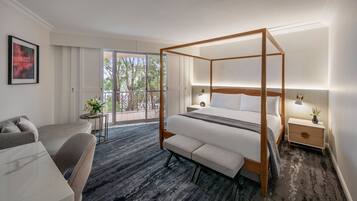 A modern hotel room with a four-poster bed, a sofa, a small table, and a balcony with a view of trees.