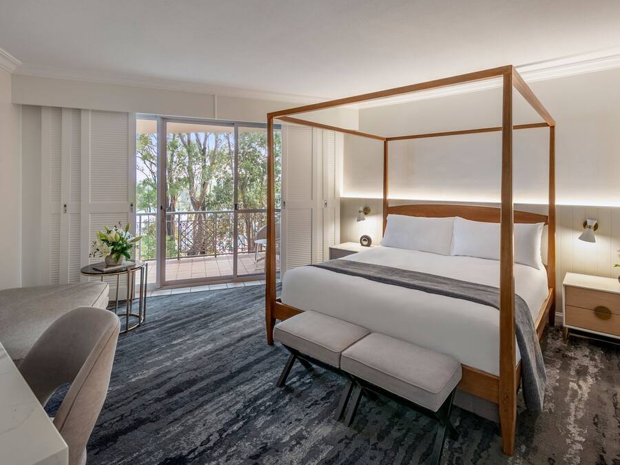 A modern hotel room with a four-poster bed, a sofa, a small table, and a balcony with a view of trees.