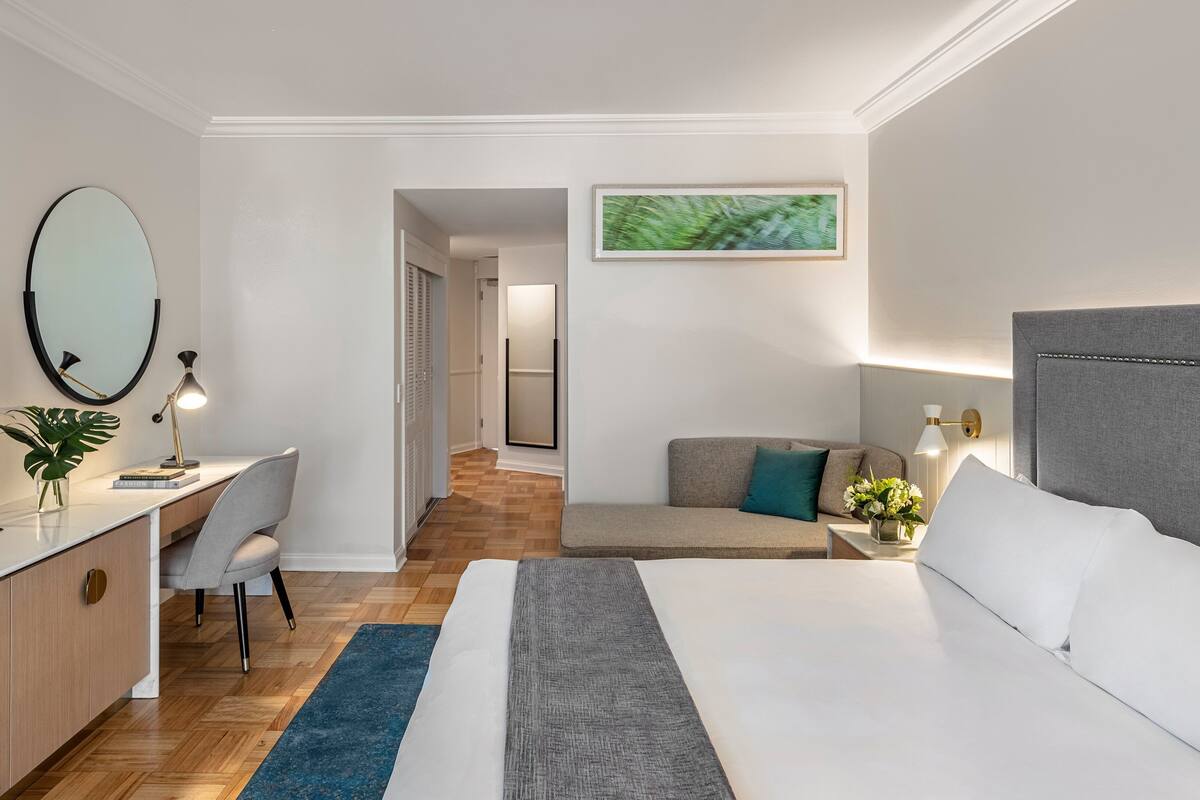 Une chambre d’hôtel comprenant un lit, un bureau, une chaise, une télévision, un miroir, un tableau encadré et une table de chevet avec une lampe.