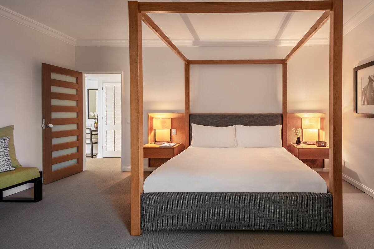Un lit à baldaquin avec un cadre en bois, deux tables de chevet avec des lampes, un fauteuil vert et un tableau encadré accroché au mur.